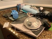 Digital white and gray Homedics noise machine on top of a white and gray device, blue rubber ice bag, black Conair hair dryer, and a Sunbeam heating pad on granite countertop under warm light.