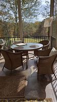 Rectangular wicker style glass top table on metal base with 4 wicker cushioned chairs on porch with wooded view in background.