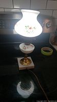 Photo of the lit 13 inch vintage table lamp showing the white glass floral hand painted shade and ceramic base with brass and marble bottom, with electrical cord.