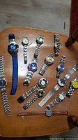 Top view of 14 wristwatches laid out on wooden surface showing variety of metal bands, rubber strap, and dial colors.