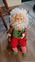Santa Claus figurine seated in wooden rocking chair, white curly hair and beard, wearing glasses, green vest, red pants, and striped socks, with power cord visible.