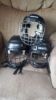 Three black kids hockey helmets with attached metal face cages, positioned on a beige fabric armchair. One helmet is Bauer brand, two are CCM brand. Straps for face cages visible.