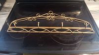 Photo of brass coloured metal coat rack placed on a stovetop, showing the entire rack with ornate scroll design and three hooks.
