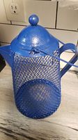 Photo of a blue metal bird feeder with mesh sides, solid lid with round knob, handle, and spout on marble countertop.