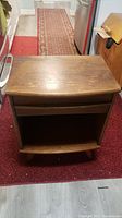 Full view of wooden side table showing top surface, drawer below, and open bottom shelf. Item placed on red carpet with partial view of rugs and kitchen appliances around.
