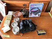 Photo shows all lot items on a wooden table including the ceramic salt pig, OptiVisor magnifier in box, black binoculars on carrying case, glass coasters with silver rims, glass ashtray, and outlet multipliers.