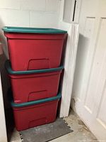 Three red storage bins with green lids stacked vertically next to a white door and insulation panels.