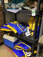 Photo of three shelves showing multiple blue and yellow life jackets, cooler, and fishing tackle box on black plastic shelving in basement setting