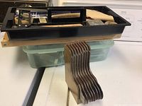 Nine L-shaped metal carving blades held vertically in a wooden holder placed on a white surface with black toolbox in background.