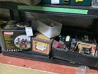 Overall view of the shelf showing Coleman heater box, wooden worm bait box, fishing reels and tackle boxes.