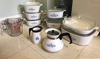 Full view of CorningWare blue cornflower cookware set with casserole dishes, glass lids, rectangular baking dishes, and FireKing white coffee pot and measuring pitcher with blue cornflower floral pattern and black handles.