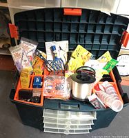 View of open plastic tackle box showing orange trays filled with assorted fishing lures, hooks, weights, bobbers, clear plastic containers, a large spool of fishing line, and a fishing reel in the main compartment.