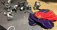 All six fishing reels laid out on a textured surface along with two cloth pouches, purple and red with gold trim.