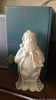 Front view of the white ceramic Lenox Santa figurine showing detailed texture and holiday greeting scroll with blue box in the background.