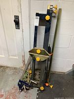 Photo showing a black and yellow miter saw stand with metal components and adjustment knobs, leaning against a white wall.