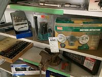 Photo showing vintage vise, drill bits, rotary tool, and boxed motion detector lamp on a shelf.