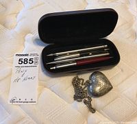 Photo of three mechanical pencils in a black case and a silverplated heart pendant with chain on a white textured surface.