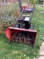 Front view of red MTD Yard Machines snow blower showing metal auger blades and black engine with controls, placed on grass.