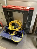 Blue metal tool box, two black and red sawhorses standing vertically behind it, yellow trouble light coiled with extension cord on top of the tool box.