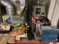 Photo showing hydraulic bottle jack box, screwdrivers, round wooden clock, and organized hardware on table with basement water heater in background.