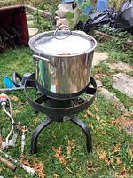 Large stainless steel pot with lid placed on cast iron burner stand outdoors on grass. Surrounding items include garden tools and debris.