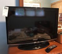 Front view of Samsung 26" TV with screen and base stand, featuring the Samsung logo on the bezel. Remote control visible beside the TV on the furniture.