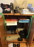 Overall view of wooden shelving unit containing various electronics, office supplies, laminator, headphones, stapler, pen set, battery charger, and AM FM weather band radio.