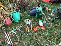 All yard and garden tools and supplies laid out on grass showing variety of items - watering cans, sprayers, shears, hand tools, soft handled garden tools.