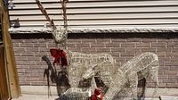 Front view of three reindeer figures standing outside against a brick wall, all with brown/red bows and woven metal wire construction with lights.