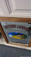Framed Southern Comfort bar sign leaning against garage door on concrete with detail of colorful house illustration and text.