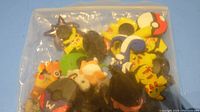 Bag of various Pokémon themed Crocs shoe charms in multiple colors and shapes visible through a plastic bag, showing several Pokémon characters and Poké Ball designs.