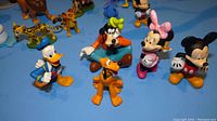 Photo showing front row of Disney character figurines including Mickey Mouse, Minnie Mouse, Goofy, Pluto, and Donald Duck on blue background.