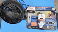 Top view photo of black electric desk fan and Kidde carbon monoxide alarm packaged in box on blue surface.