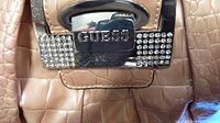 Close-up of the Guess logo on the purse with rhinestone detailing on the clasp in tan crocodile texture material.