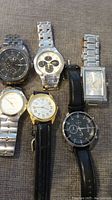 Six men's wristwatches laid out on fabric surface showing variety of styles and bands.