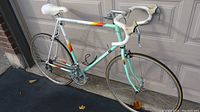 Left side view of vintage Peugeot PS28 men's road bike. Shows light turquoise and white frame with orange/yellow decals, drop handlebars with white tape, white saddle, and thin 700c tires.