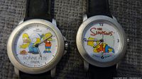 Two wristwatches side by side, one with Homer and Bart Simpson characters and French text on dial, the other with Bart Simpson reclining and The Simpsons logo.