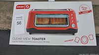 Photo of Dash Clear View Toaster box showing red toaster with visible clear window and control knob, text describing features including 7 level browning control and 1100 watts.