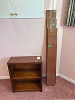 Photo showing small wooden bookshelf with two shelves next to five wooden shelf boards leaning against the wall