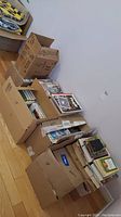 Eight cardboard boxes packed with assorted books and magazines, stacked on a wooden floor against a wall.