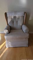 Front view of wing chair showing patterned fabric upholstery, doilie style cover on top backrest, padded arms with arm covers, and skirted bottom.