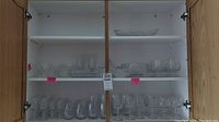 Full view of two shelves in a glass cabinet with different crystal glassware items. Mostly clear glasses and some dishes, arranged side by side.