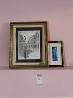 Two framed art prints displayed on a pink wall, one larger with ornate gold frame, the other smaller in a dark frame.