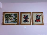 Three framed needlepoint pieces displayed on a wall: a colorful street scene and two dog portraits with hats and clothing.