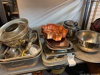 Group of various metal baking pans, molds, and trays stacked together including a copper-tone flower-shaped mold and a large stainless steel mixing bowl.