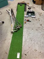 Overview of the golf clubs aranged on a green putting mat along with two putting machines and golf accessories such as balls and tees.