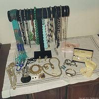 Front view of assorted jewelry pieces including necklaces of various colors on a black velvet T-bar jewelry holder. Several bracelets, earrings, and rings are laid out on a white cloth underneath.