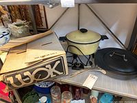 Vintage cream-colored electric pot with glass lid and black metal stand, and a large black fry pan with lid on a shelf next to the box labeled 'Wok'.