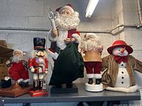 Photo showing five Christmas figurines: four Santas and a nutcracker on top of a metal shelf against a gray cinder block wall under a fluorescent light.