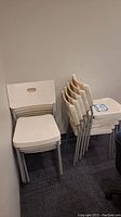 Stack of 10 white Ikea chairs with metal legs in indoor setting against grey carpeted floor and plain wall.